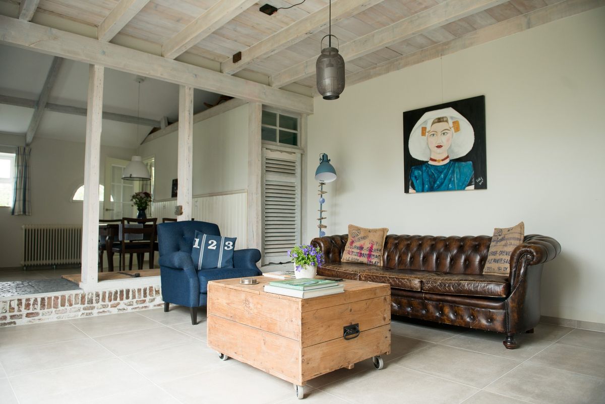 Woonkamer met leren Chesterfield bank, blauwe fauteuil en houten balken – Zeeuwshuisje vakantiewoning, Koudekerke